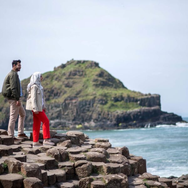 2 people on top of the rock formations, with sea and distant shoreline in view.
