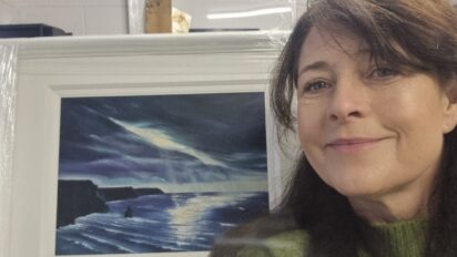 Ann Daly posing beside her framed painting. A painting of the Cliffs of Moher in Ireland, a mixture of blues and greys.